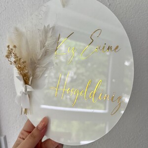 May include: White round acrylic sign with gold lettering that reads "Kız Evine Hosgeldiniz". The sign is decorated with dried white and beige flowers.