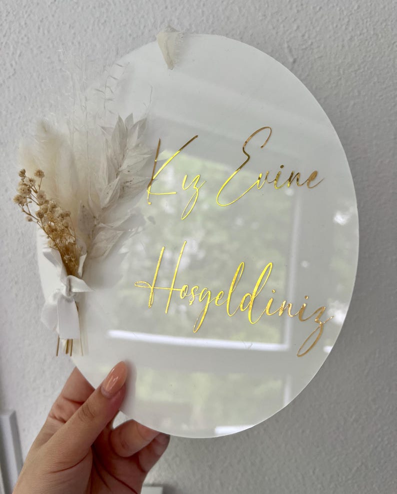 May include: White round acrylic sign with gold lettering that reads "Kız Evine Hosgeldiniz". The sign is decorated with dried white and beige flowers.