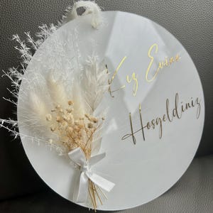 May include: A white round sign with gold lettering that says "Welcome" in Turkish. The sign is decorated with dried white pampas grass and a small bouquet of dried white and beige flowers tied with a white ribbon.