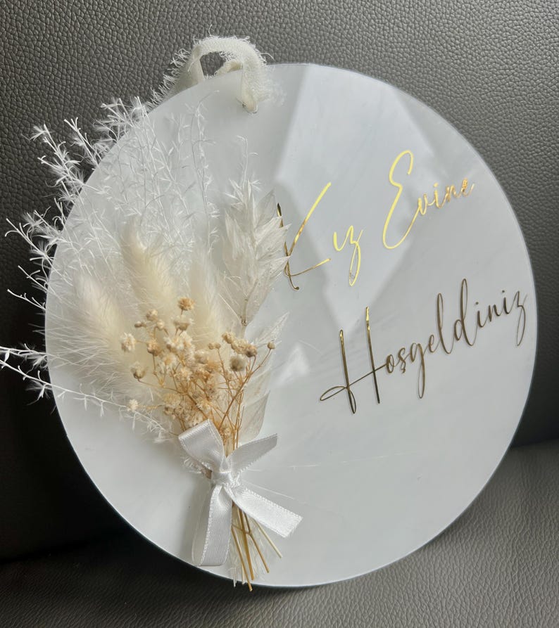 May include: A white round sign with gold lettering that says "Welcome" in Turkish. The sign is decorated with dried white pampas grass and a small bouquet of dried white and beige flowers tied with a white ribbon.