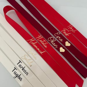 Könnte beinhalten: Sechs weiße Satinbänder mit schwarzer Beschriftung und zwei rote Satinbänder mit goldener Beschriftung. Die weißen Bänder tragen die Namen "Tarkan", "Tugba", "Selin", "Selin", "Mohammed" und "Natalia". Die roten Bänder tragen die Namen "Esra", "Okan" und "Nagih Faik" mit einem goldenen Herzsymbol.