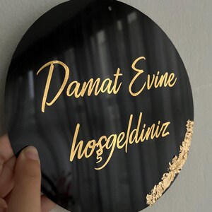 May include: A black circular sign with gold lettering that reads "Damat Evine hosgeldiniz". The sign has a gold accent on the right side.