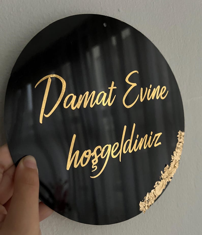 May include: A black circular sign with gold lettering that reads "Damat Evine hosgeldiniz". The sign has a gold accent on the right side.