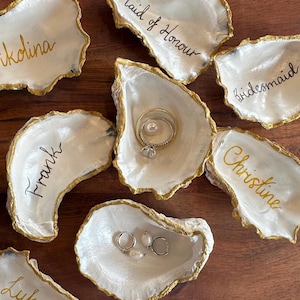May include: A collection of oyster shell ring dishes, each with gold edging and handwritten names. One shell holds a ring with a pearl and diamond. Other shells are inscribed with names such as "Maid of Honour" and "Bridesmaid".