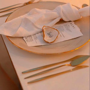 May include: A wedding table setting with a beige plate with a gold rim, a white linen napkin, and gold cutlery. An oyster shell with the word "Bride" is placed on a menu card. The scene is illuminated with soft, natural light.