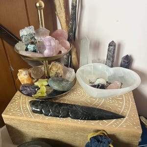 May include: A two-tiered gold stand with a variety of crystals and stones, including pink, purple, green, and clear. The stand is on a wooden box with an intricate carved design. A heart-shaped selenite bowl with crystals and a black obsidian dagger are also on the box.