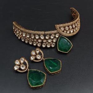 May include: A gold choker necklace with a green gemstone pendant and matching earrings. The necklace is decorated with clear stones and the earrings have a floral design.
