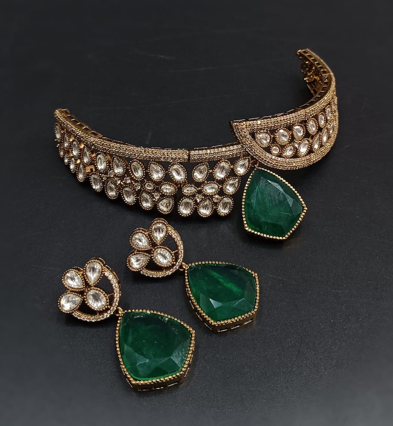 May include: A gold choker necklace with a green gemstone pendant and matching earrings. The necklace is decorated with clear stones and the earrings have a floral design.