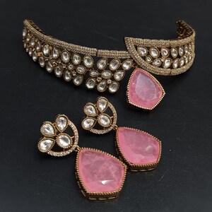 May include: A gold choker necklace with a pink stone pendant and matching earrings. The necklace is decorated with clear stones and the earrings have a floral design.