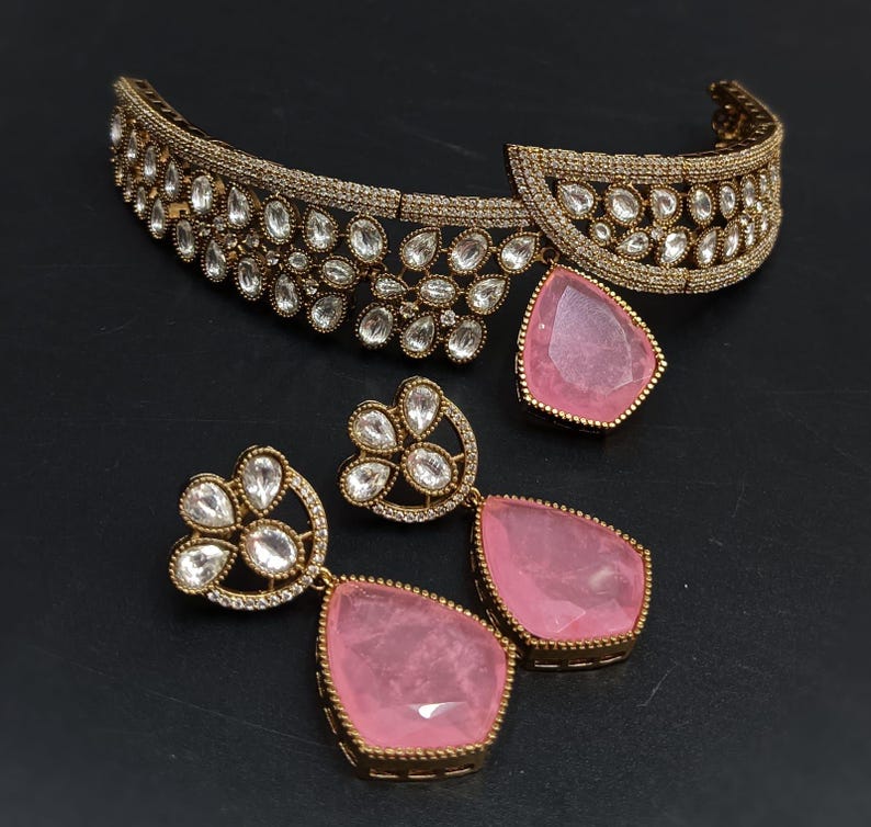 May include: A gold choker necklace with a pink stone pendant and matching earrings. The necklace is decorated with clear stones and the earrings have a floral design.