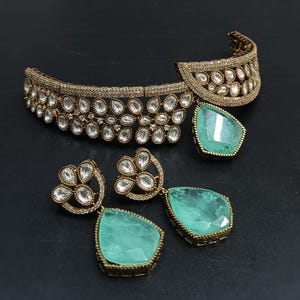 May include: A gold choker necklace with a teardrop-shaped green stone pendant and matching earrings. The necklace is decorated with clear stones in a floral pattern.