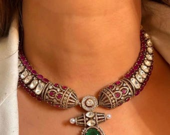 Op de afbeelding: Een sierlijke ketting met een grote smaragdgroene hanger. De ketting heeft een gebogen ontwerp met robijn- en diamantaccenten. De hanger is omgeven door een halo van kleinere diamanten en een parel.