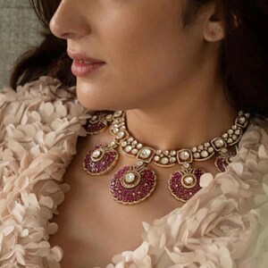 May include: A woman wearing a cream-colored, ruffled top with a gold and diamond necklace featuring large ruby accents.