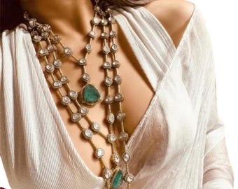 May include: A layered gold necklace with emerald green gemstones and diamond accents. The necklace is worn by a person with long dark hair and a white top.