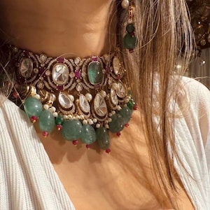 Puede incluir: Un collar gargantilla de oro con una gran piedra preciosa verde esmeralda, rodeada de diamantes más pequeños y piedras rubí rojas. El collar está adornado con una franja de perlas verde esmeralda y blancas.