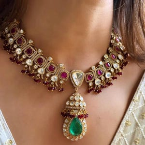 May include: A gold necklace with a large green gemstone pendant. The necklace is decorated with small red and white stones.
