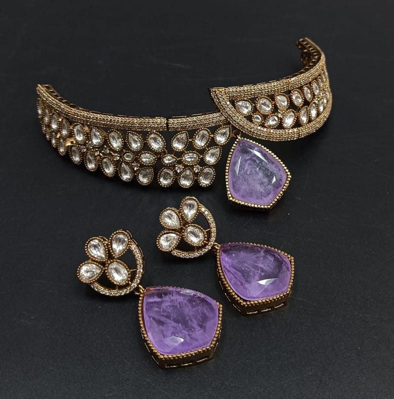 May include: A gold choker necklace with a purple gemstone pendant and matching earrings. The necklace is decorated with clear stones and the earrings have a floral design.