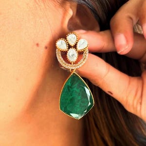 May include: Gold dangle earrings with a teardrop-shaped green gemstone and a cluster of white stones in a floral design.