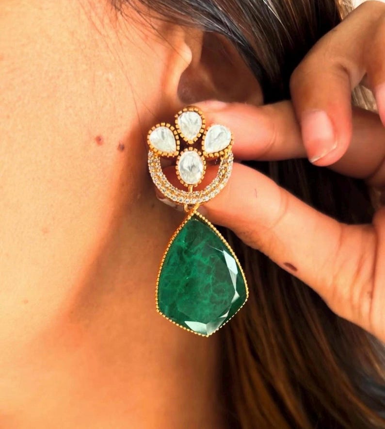 May include: Gold dangle earrings with a teardrop-shaped green gemstone and a cluster of white stones in a floral design.