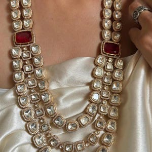May include: A multi-strand gold necklace featuring red and clear stones. The necklace is crafted from multiple layers of gold chains with large, square-shaped stones.