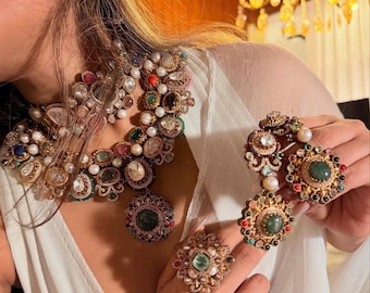 May include: A gold-toned jewellery set comprising a necklace, earrings, a bracelet, and rings. The pieces are embellished with colourful gemstones, pearls, and detailed designs. The necklace and rings feature large, central green stones.