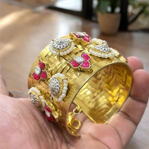 May include: A wide gold-coloured cuff bracelet with a woven design. The bracelet is adorned with silver and pearl accents, and red gemstones. The bracelet is held in a hand.