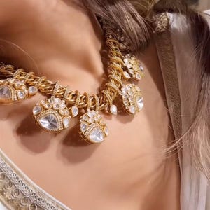 May include: A gold necklace with large, clear, teardrop-shaped pendants. The necklace features a thick chain design with smaller, clear stones. The necklace is worn over a white garment with gold trim.