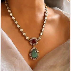 May include: A gold necklace with a teardrop-shaped pendant featuring a green stone surrounded by smaller white stones. The pendant is attached to a chain of white stones.