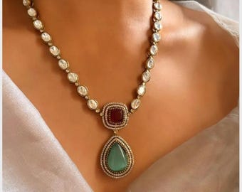 May include: A gold necklace with a teardrop-shaped pendant featuring a green stone surrounded by smaller white stones. The pendant is attached to a chain of white stones.