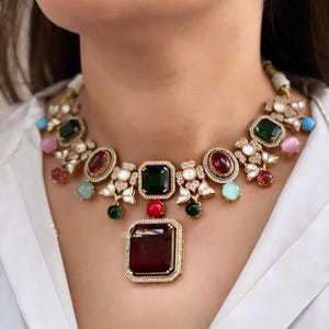 Może przedstawiać: Złoty naszyjnik z dużym czerwonym kamieniem szlachetnym wisiorkiem otoczonym mniejszymi kamieniami szlachetnymi w różnych kolorach, w tym zielonym, niebieskim i różowym.