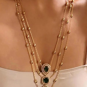 Pode incluir: Um colar de corrente dourada em camadas com um pingente com três pedras de esmeralda verde cercadas por pedras de zircônia cúbica transparentes.