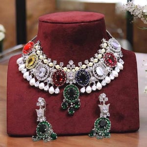 Peut inclure: Un collier argenté avec un motif de pierres multicolores. Le collier présente une rangée de pierres rondes en rouge, jaune, vert, bleu et violet, entourées de petites pierres blanches. Les boucles d'oreilles sont de conception assortie avec une seule pierre verte.
