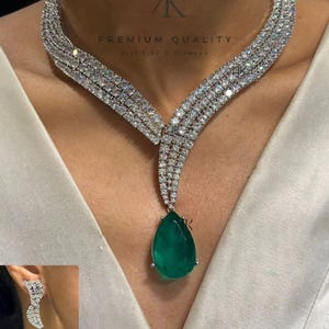 May include: A silver necklace with a large emerald green pear-shaped pendant. The necklace is made of multiple rows of small, clear stones. The pendant is surrounded by smaller clear stones. The necklace is paired with matching earrings with a similar design.