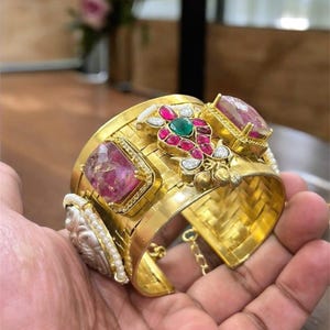 May include: A wide, gold-coloured cuff bracelet with intricate detailing. The bracelet features large, rectangular pink gemstones, smaller red and green stones, and white accents. The design includes a woven texture and a small chain.