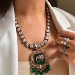May include: A gold necklace with a green and white pendant featuring an elephant design. The pendant is surrounded by small white stones and green beads.