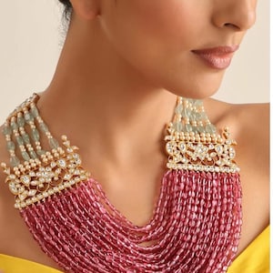 Op de afbeelding: Een meerrijige ketting met een gouden en witte hanger. De ketting is gemaakt van roze en groene kralen.