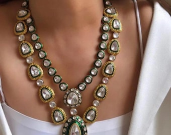 Op de afbeelding: Een ketting met twee strengen met een goudkleurige zetting, met grote, heldere, druppelvormige stenen en kleinere, ronde stenen. De ketting heeft groene en gouden accenten en een grote hanger aan de onderkant.