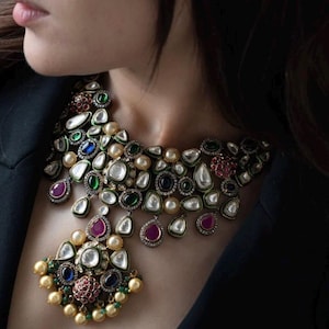 Puede incluir: Un collar de plata y oro con un gran colgante. El collar está adornado con perlas, esmeraldas, rubíes y zafiros. El colgante tiene forma de lágrima y presenta un racimo de perlas y piedras preciosas.