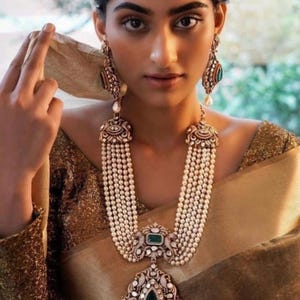 May include: A woman wearing a gold and white saree with a long, multi-strand pearl necklace with a large, ornate pendant featuring a green gemstone. The pendant is decorated with intricate details and smaller gemstones. She is also wearing matching earrings with green gemstones.