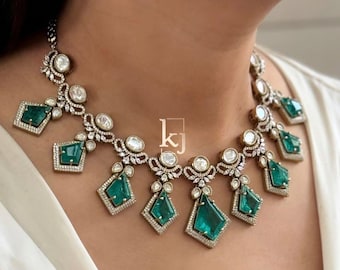 May include: A silver and emerald green statement necklace with a delicate design. The necklace features a central pendant with a large emerald green stone surrounded by smaller diamonds. The pendant is connected to a chain with smaller emerald green stones and diamonds.