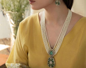 May include: A woman wearing a yellow top with a long pearl necklace and a green and gold pendant. She is also wearing green and gold earrings.