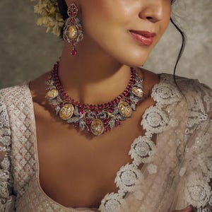 May include: A close-up of a woman wearing a necklace and earrings. The necklace is made of gold and has red and white stones. The earrings are also made of gold and have red stones.