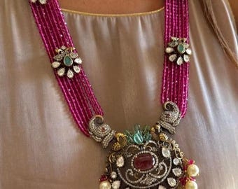 May include: A long, layered necklace with a large, ornate pendant. The necklace is made of pink beads and features a silver pendant with a red gemstone and white pearls. The pendant is decorated with intricate details and is a beautiful example of traditional Indian jewelry.