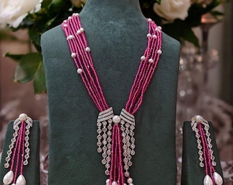 May include: A magenta beaded necklace and matching earrings set displayed on a green velvet bust. The necklace features multiple strands of magenta beads, accented with white pearls and diamond-like embellishments. The earrings have a similar design.