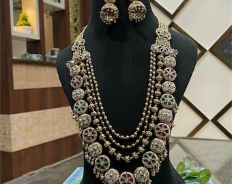 May include: A gold-toned necklace and earring set displayed on a black mannequin. The necklace features multiple strands of small beads and ornate, circular pendants with red, green, and white accents. The earrings mirror the pendant design. The jewelry is likely for special occasions.