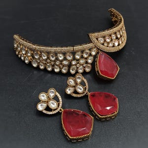 May include: A gold-tone choker necklace with a red gemstone pendant and matching earrings. The necklace and earrings are adorned with clear cubic zirconia stones.