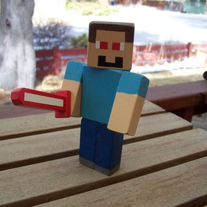 Może przedstawiać: Drewniana figurka postaci z Minecrafta trzymająca czerwony miecz. Figurka ma niebieską koszulę, niebieskie spodnie i brązową głowę z czerwonymi oczami.