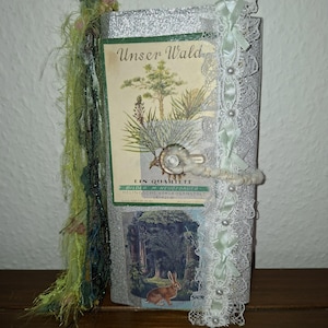 Könnte beinhalten: Ein handgefertigtes Journal mit Vintage-Ästhetik. Das Cover zeigt eine botanische Illustration mit dem Text "Unser Wald". Verzierungen sind Spitze, Bänder und ein dekorativer Verschluss. Der Buchrücken hat grüne und schwarze Fransen.