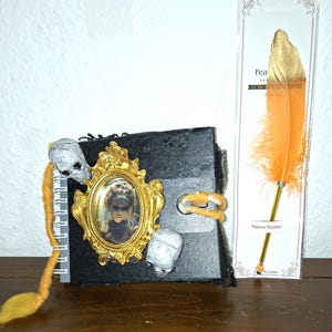 Op de afbeelding: Een zwart notitieboek met een goudomrand portret en schedelversieringen. Een decoratieve oranje veren pen met gouden accenten wordt naast het notitieboek weergegeven. Het notitieboek heeft een spiraalbinding en een sluiting.