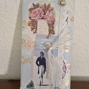 Könnte beinhalten: Ein hellblaues Notizbuch mit floralen und Vintage-Illustrationen. Das Cover zeigt einen rosa Rosenstrauß, ein Porträt eines Mannes und den Text "Mr. Darcy & Elizabeth Bennet". Verziert mit Spitze und einem dekorativen Juwel.
